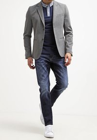 Grå kavaj över en marinblå polo-shirt, ihop med blå jeans och vita sneakers. Slät tyg, skräddarsydd design och avslappnad passform.