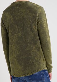 Olivgrön långärmad skjorta med en texturerad, blekt utseende. Har en rund halsringning och stickad fåll. Bärs tillsammans med blå jeans.