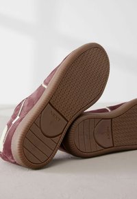 Semelles de baskets en daim marron avec semelle en caoutchouc à crampons, marque "NEXT" et taille 40 visibles en dessous, positionnées sur un fond uni.