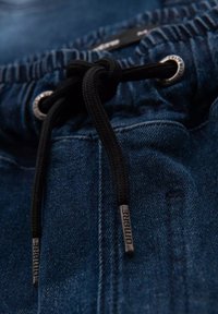 Joggers in denim blu scuro con vita regolabile tramite coulisse, caratterizzati da cordoni neri intrecciati e occhielli metallici. La vista ravvicinata mette in evidenza la texture e le cuciture.