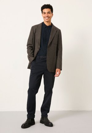 Junger Mann lächelt, trägt einen dunkel karierten Blazer, schwarzes Hemd mit Knöpfen, schwarze Hose und schwarze Stiefel, steht vor hellem Hintergrund.