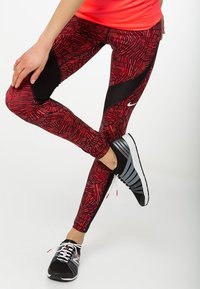 Czerwone legginsy sportowe w wzory z czarnymi siateczkowymi akcentami; w zestawie z czarno-szarymi butami do biegania z różowymi detalami i białymi sznurówkami.