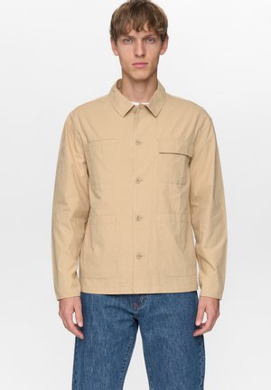 Jonge man die een beige jas met knopen en drie voorzakken draagt, met een blauwe spijkerbroek, staand tegen een effen witte achtergrond.