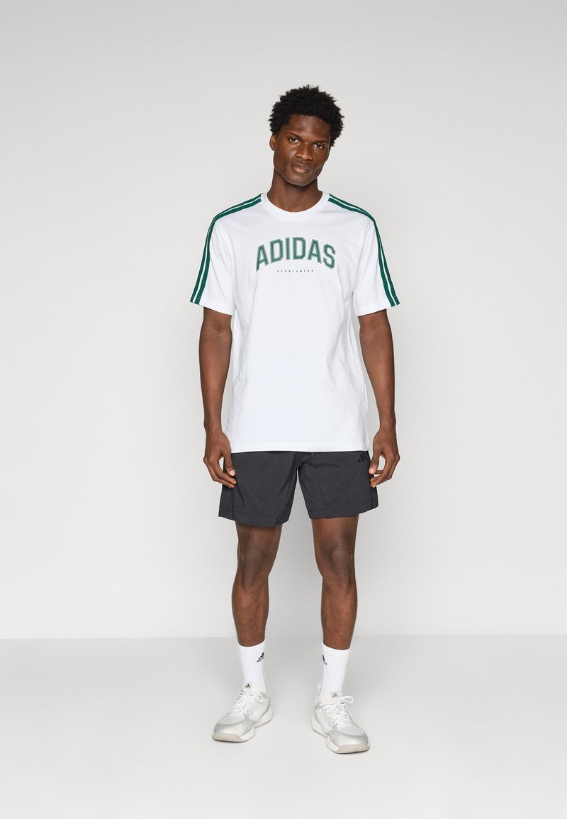 Witte t-shirt met korte mouwen met groene tekst "ADIDAS" en drie groene strepen op de schouders, gecombineerd met zwarte short en witte sneakers.