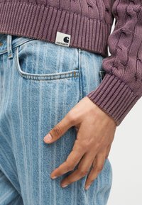 Χέρι που ακουμπά στην τσέπη ανοιχτομπλε τζιν, φορώντας μοβ πουλόβερ με πλεκτό σχέδιο και εμφανή ετικέτα λογότυπου Carhartt στον ποδόγυρο.
