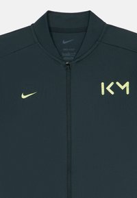 Μαύρη αθλητική ζακέτα Nike Dri-FIT με φερμουάρ, με κίτρινο λογότυπο "KM" στο δεξί στήθος και κίτρινο σήμα Nike στο αριστερό στήθος.