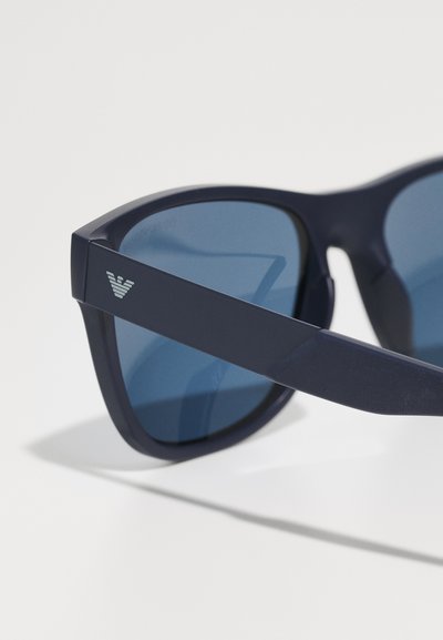 Ochelari de soare navy cu lentile de culoare albastru închis și finisaj mat. Prezintă o formă de cadru triunghiulară și un logo argintiu pe bradă.