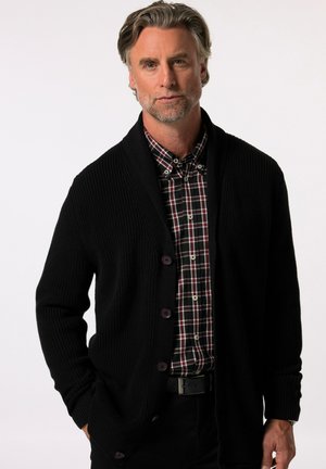 Cárdigan negro de punto con cuello de chal y cierre de botones, superpuesto sobre una camisa a cuadros en rojo, negro y blanco. Textura suave en todo.