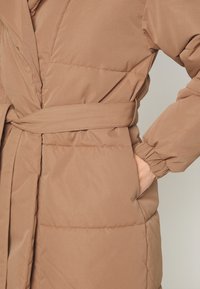 Hnědý prošívaný kabát s opaskem, bočními kapsami a elastickými manžetami. Tkanina je hladká s jemnou texturou a prošívanými panely.