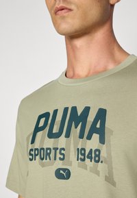 Maslinasto zelena pamučna majica s okruglim izrezom, s istaknutim tamnozelenim grafikonom "PUMA SPORTS 1948" u reljefnom printu.