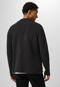 Svart texturerad jacka med krage, lång ärm och en rak bakseam, bärs över en vit skjorta. Materialet verkar tjockt och varmt.