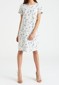 Frau trägt ein weißes knielanges Kleid mit kurzen Ärmeln und blauem Blumenmuster, kombiniert mit nudefarbenen, ankle-tie High-Heels.