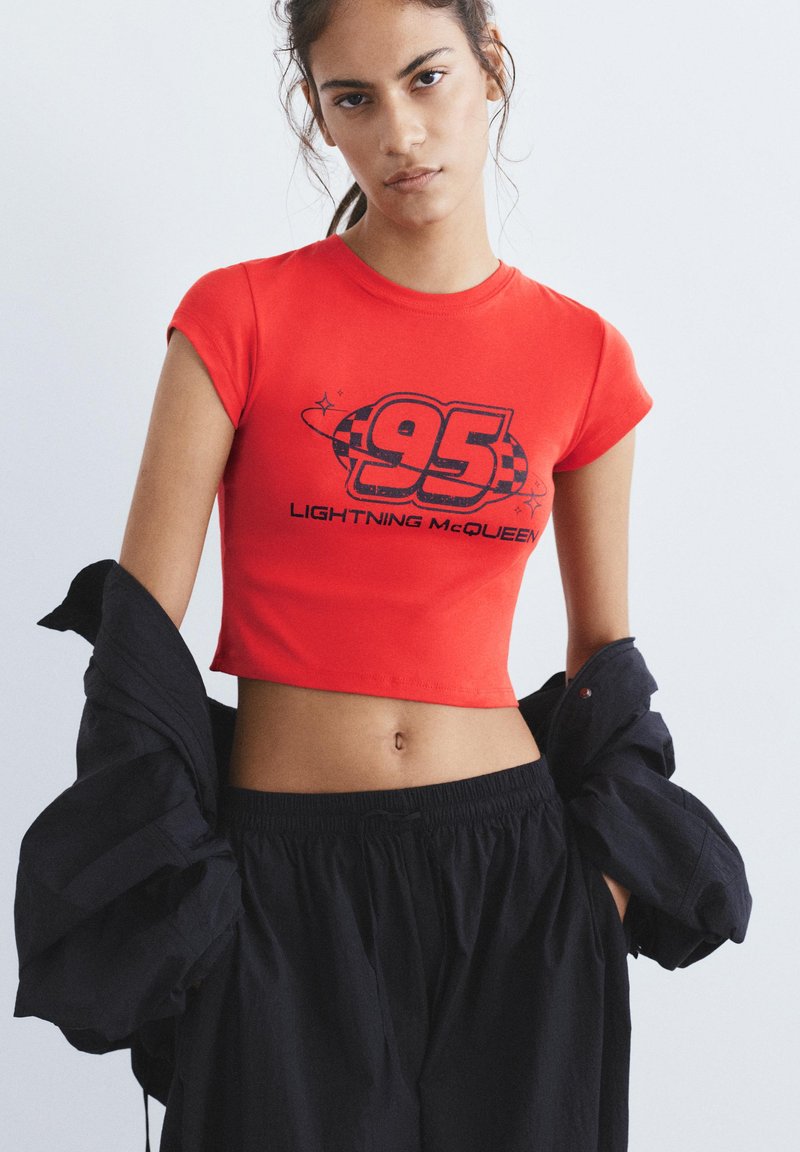 Jonge vrouw draagt een rood crop-topje met de tekst '95 Lightning McQueen' en zwarte broek, houdt een zwart jasje rond haar ellebogen.