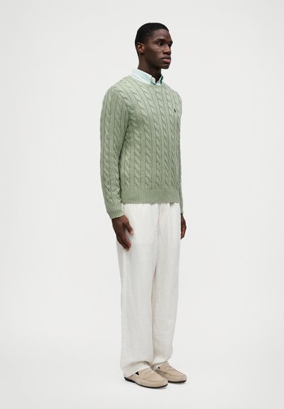 Ανοιχτό πράσινο πουλόβερ με πλέξη, λευκό πουκάμισο από λινό παντελόνι και μπεζ παπούτσια. Το σύνολο διαθέτει πουκάμισο με γιακά κάτω από το πουλόβερ.