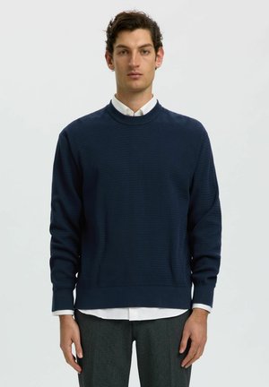 Uomo che indossa un maglione blu navy strutturato sopra una camicia bianca e pantaloni grigi, in piedi davanti a uno sfondo chiaro e uniforme.