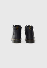 Botas de tornozelo pretas com acabamento em couro liso, colarinho acolchoado, fechos de correr na parte traseira e etiqueta da marca amarela. Apresenta uma sola de borracha robusta com detalhes em costura.
