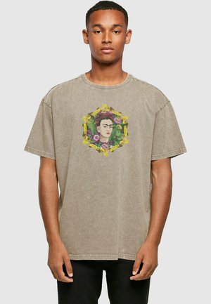 Lys brune bomulds t-shirt med et farverigt grafik af en kvinde omgivet af blomster og bånd i et udsmykket design. Korte ærmer, løs pasform.