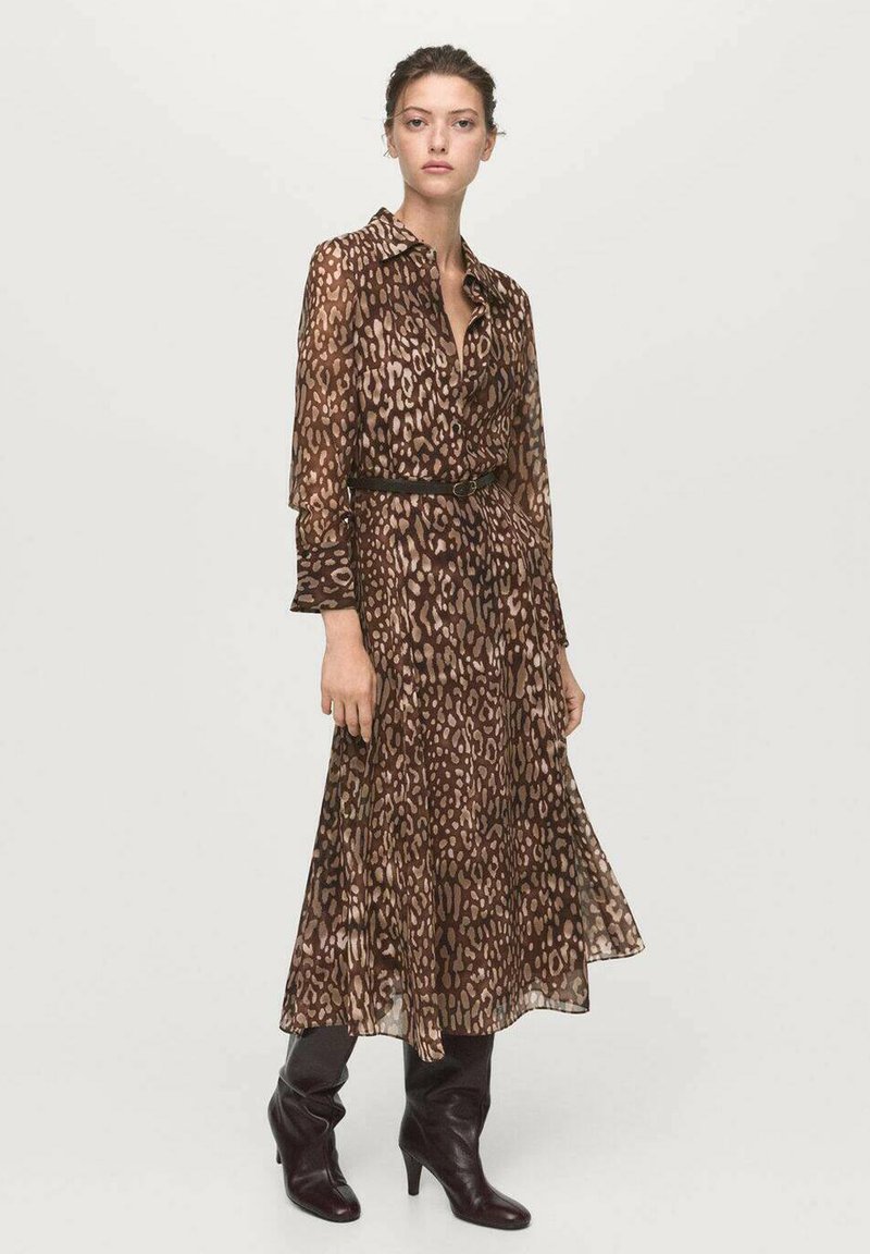 Mujer vestida con un vestido midi de manga larga de estampado de leopardo marrón, con un cinturón negro y botas de tacón negras, de pie sobre un fondo liso.
