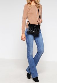 Borsa a tracolla nera in ecopelle con texture a coccodrillo, forma rettangolare, tracolla regolabile e patta frontale. Indossata con cardigan beige e jeans.