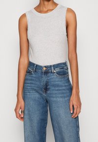 Top sin mangas de color gris claro con escote redondo, combinado con jeans azules de cintura alta que tienen un cierre de botón y bolsillos frontales.