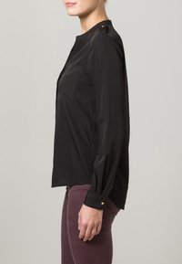 Blusa nera a maniche lunghe con colletto alla cinese, tasca anteriore e dettagli in oro sui bottoni delle spalle. Tessuto morbido e design aderente.