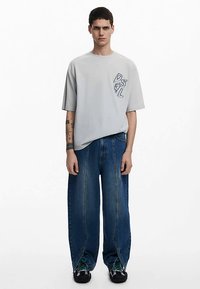 Grijze oversized T-shirt met geborduurd logo, gecombineerd met ruime blauwe spijkerbroek met een gespleten zoom en zwarte schoenen.