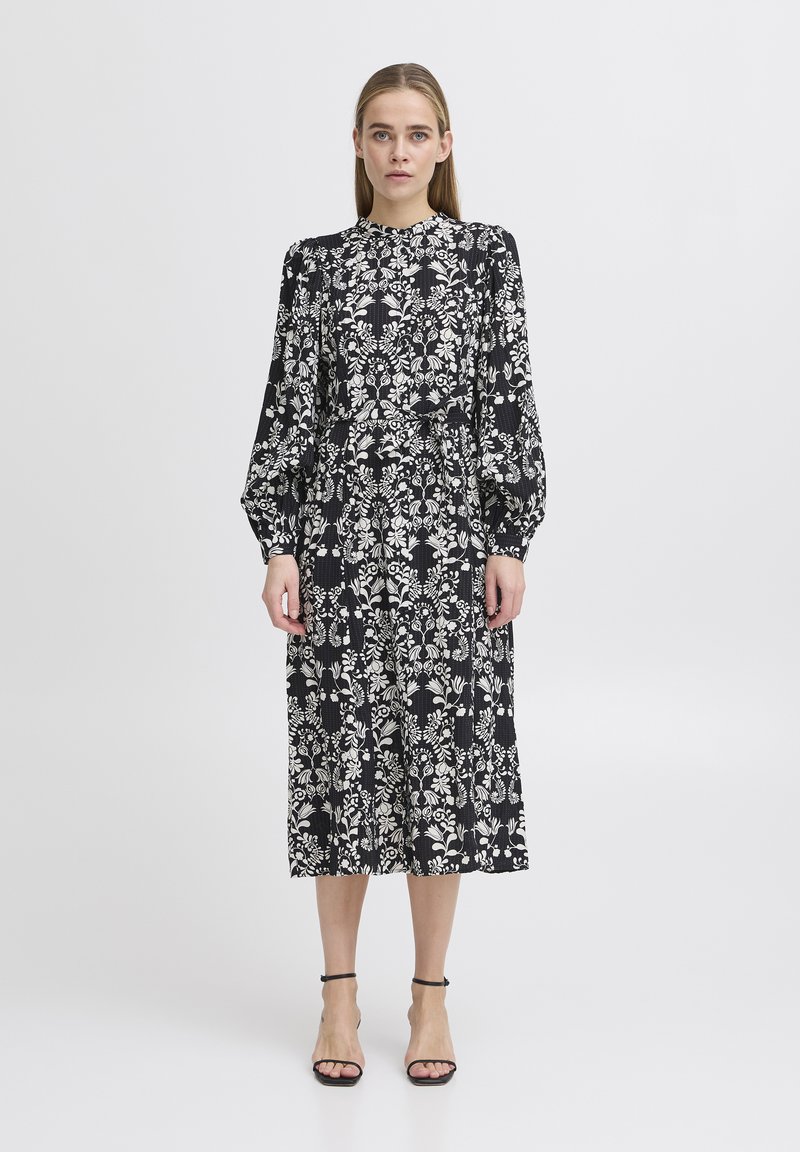 Vestido midi negro con patrones florales blancos, mangas largas abullonadas, escote alto y cinturilla ceñida, combinado con tacones negros con tiras.