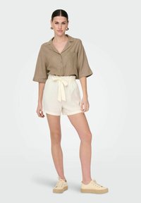 Camicetta marrone a maniche corte con bottoni e tasca, abbinata a pantaloni corti bianchi alti in vita con un fiocco. Scarpe beige con lacci.