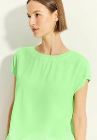 Top a maniche corte in tessuto verde brillante con scollatura rotonda e dettagli arricciati lungo il collo. Texture liscia e silhouette casual.
