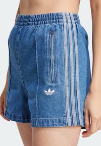 Pantalones cortos de mezclilla azul con cinturilla elástica, bolsillo lateral con cremallera y tres franjas blancas a los lados. Presenta un pequeño logo bordado.
