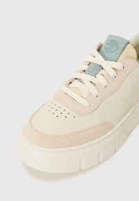 Beige lærersneaker med en teksturert semsket overflate, kremfargede lisser og en lyseblå aksent på tungen med logo. Perforert tåområde.