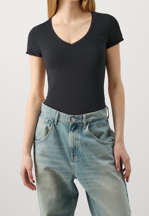 Femme portant un t-shirt noir ajusté à manches courtes en V rentré dans un jean ample bleu clair taille haute, sur un fond uni.
