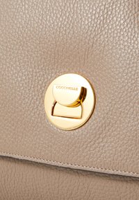 Beige textured leather handbag featuring a circular gold hardware clasp with "Coccinelle" engraved. Smooth stitching along the edges.
