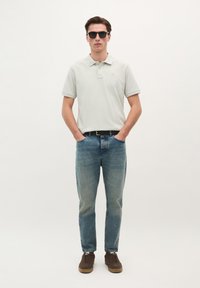 Helles graues Poloshirt mit kurzen Ärmeln, blaue Jeans, braune Schuhe und dunkle Sonnenbrille, stehend vor einem schlichten Hintergrund.