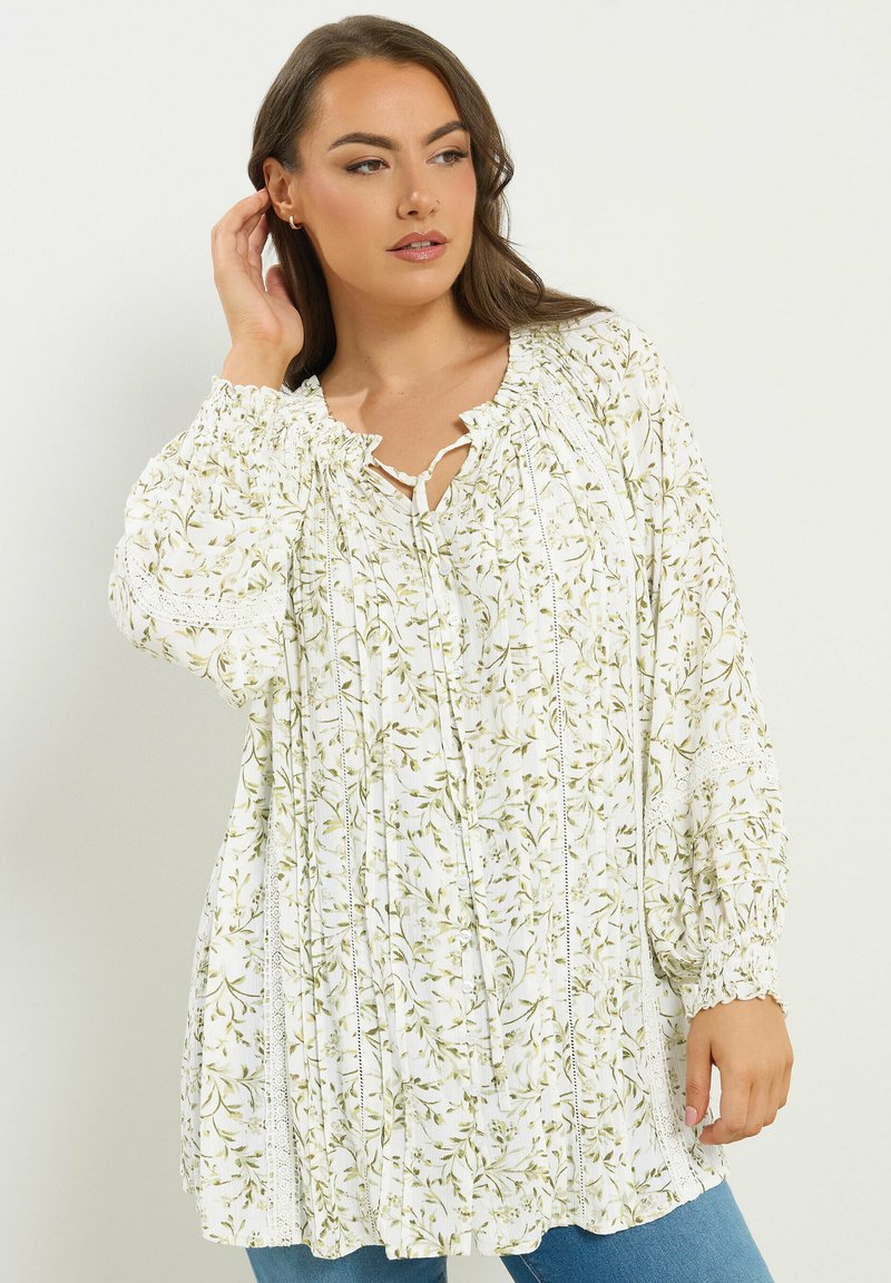 Vrouw in een losse witte blouse met een groen bloemenpatroon en kanten details, poserend met één hand die haar oor raakt en zijwaarts kijkt.