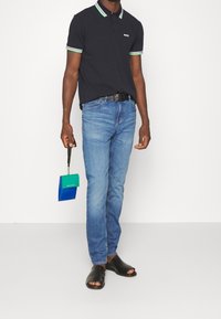 Camisa polo negra con cuello a rayas verdes y blancas, jeans de mezclilla azules, sandalias negras y un bolso pequeño bicolor sostenido con la mano izquierda.