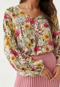 Blusa floreale a maniche lunghe con scollo a V, caratterizzata da motivi di fiori rosa, gialli e verdi su sfondo crema, abbinata a una gonna plissettata rosa.