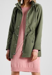 Veste utilitaire vert olive avec taille ajustable par cordon, fermetures par boutons-pression et capuche, portée sur une robe en maille rose avec ourlet côtelé.
