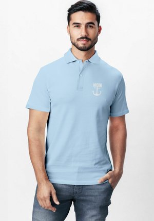 Helles blaues Poloshirt aus Baumwolle, mit Kragen und kurzen Ärmeln. Es hat auf der Brust einen "MOIN"-Text und ein Anker-Graphic in hellen Farben.