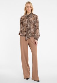 Woman wearing a leopard-print sheer blouse with a tie neck and tan wide-leg pants, standing with one hand in pocket against a white background.