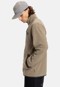 Fleece jacket in tan with a zippered pocket, black accents, side pockets, and a stand collar. Paired with a gray cap.