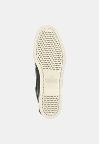Zapato negro con suela de goma que presenta un patrón de banda de rodadura ondulado. Parte superior texturizada con un diseño casual. La suela es de color crema con un ligero branding.