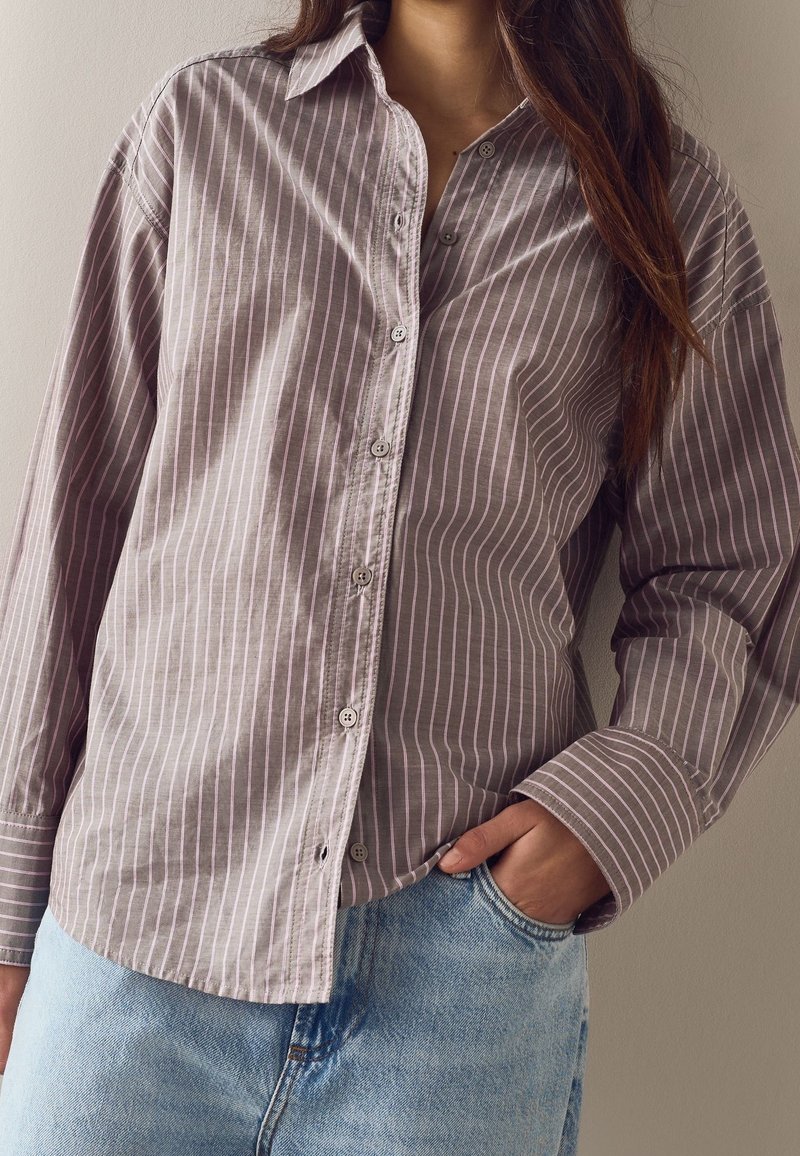 Personne portant une chemise à manches longues rayée violet clair, rentrée dans un jean bleu clair, avec une main dans la poche.