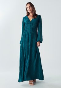 Žena v tyrkysových dlouhých rukávech, zmačkaných maxi šatech s knoflíky vpředu, stojící proti jednoduchému světelnému pozadí.