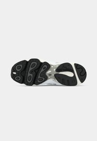 Sort og grå mønstret gummisål på en New Balance sneakers set fra neden på en hvid baggrund.