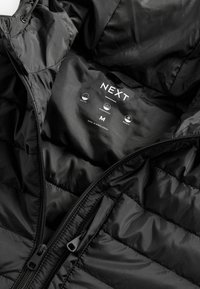 Černá prošívaná bunda se zipem a kapucí. Vnitřní štítek ukazuje značku "NEXT", velikost "M" a ikony pro prošívání, voděodolnost a tepelné vlastnosti.