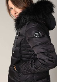 Doudoune noire avec une capuche en fausse fourrure, texture matelassée et patch logo sur la manche. Les caractéristiques comprennent des poches zippées et un réglage de la capuche à cordon.