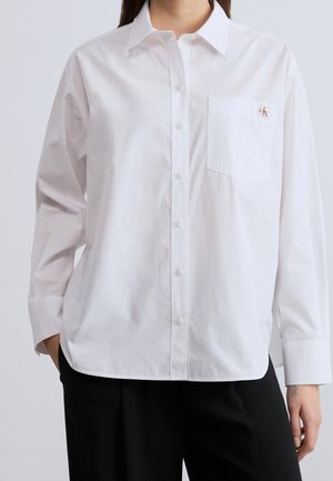 Vrouw draagt een wit overhemd met knoopjes en een voorzak met een klein rood "cK"-logo, gecombineerd met zwarte broek.