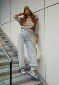 Ljust grå höga vida jeans, beige och brun färgblockerad hoodie, grå och vita Nike-sneakers, bärande en texturerad vit väska.