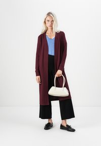 Bordeaux lange cardigan over een blauwe v-hals top, gecombineerd met zwarte wijde broek en zwarte schoenen, terwijl ze een witte handtas vasthoudt.